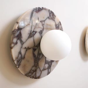 Pode incluir: Uma lumin&aacute;ria de parede moderna com uma base de m&aacute;rmore branco com veios cinzentos, combinada com uma luz branca esf&eacute;rica. Outra lumin&aacute;ria com um detalhe em madeira &eacute; vis&iacute;vel no fundo.