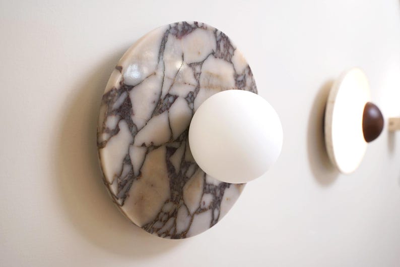 Pode incluir: Uma lumin&aacute;ria de parede moderna com uma base de m&aacute;rmore branco com veios cinzentos, combinada com uma luz branca esf&eacute;rica. Outra lumin&aacute;ria com um detalhe em madeira &eacute; vis&iacute;vel no fundo.