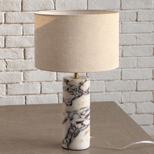 May include: A table lamp featuring a cylindrical base made of white marble with prominent dark purple veining. The lamp is topped with a neutral-toned, drum-shaped fabric shade and includes a thin gold-colored metal connector piece.