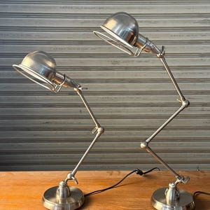 May include: Two silver-toned desk lamps with adjustable arms and round bases. Each lamp features a domed shade and a wire guard. The lamps are positioned on a wooden surface, with a neutral background.