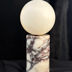 May include: A table lamp featuring a cylindrical base made of white and deep purple veined marble. The base supports a spherical, off-white glass globe shade. The lamp has a smooth, polished finish and a vertical orientation, standing upright.