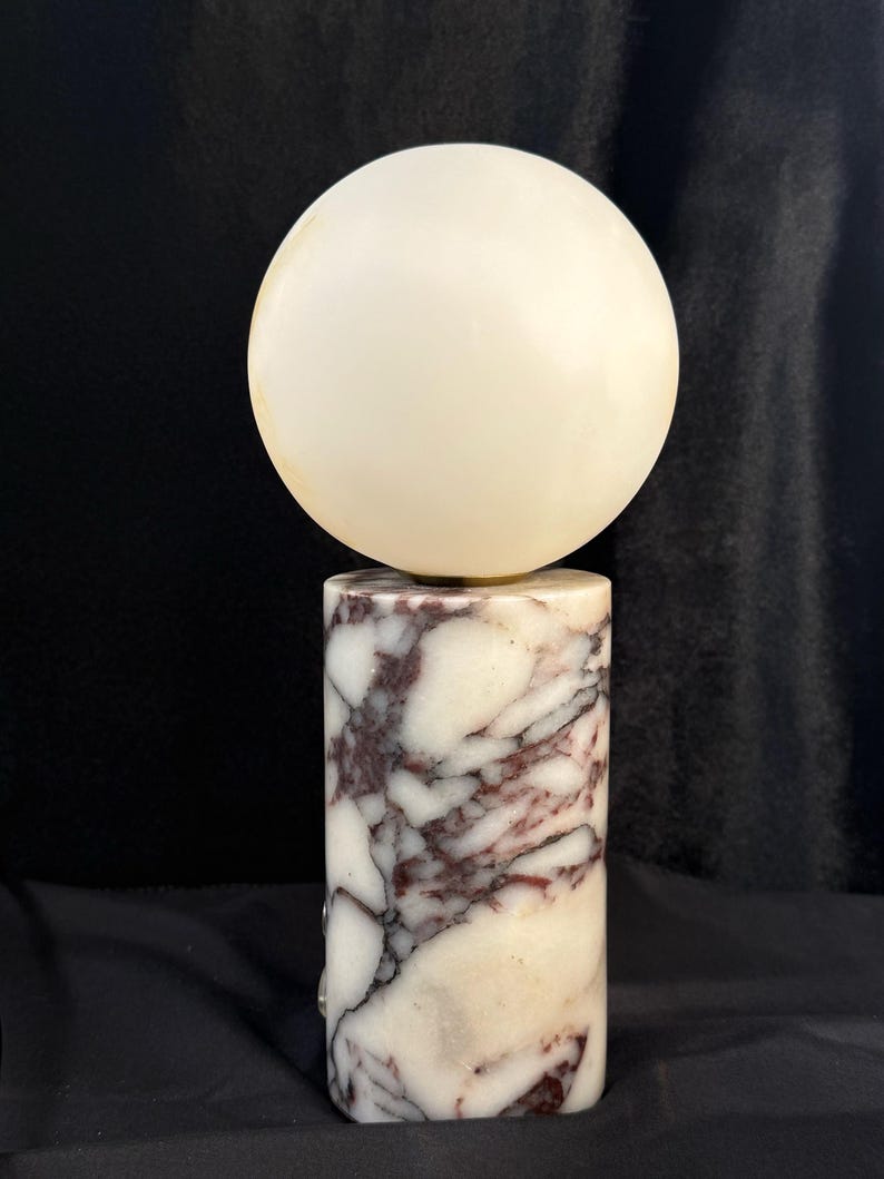 May include: A table lamp featuring a cylindrical base made of white and deep purple veined marble. The base supports a spherical, off-white glass globe shade. The lamp has a smooth, polished finish and a vertical orientation, standing upright.