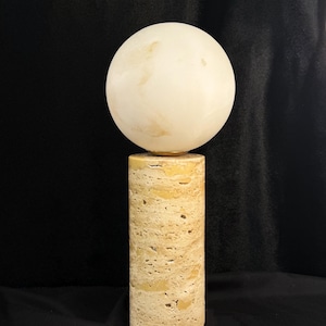 May include: A table lamp featuring a cylindrical base made of porous, beige travertine stone. The base supports a smooth, spherical, off-white globe shade that sits directly on top of the stone column, creating a balanced and geometric lighting fixture.
