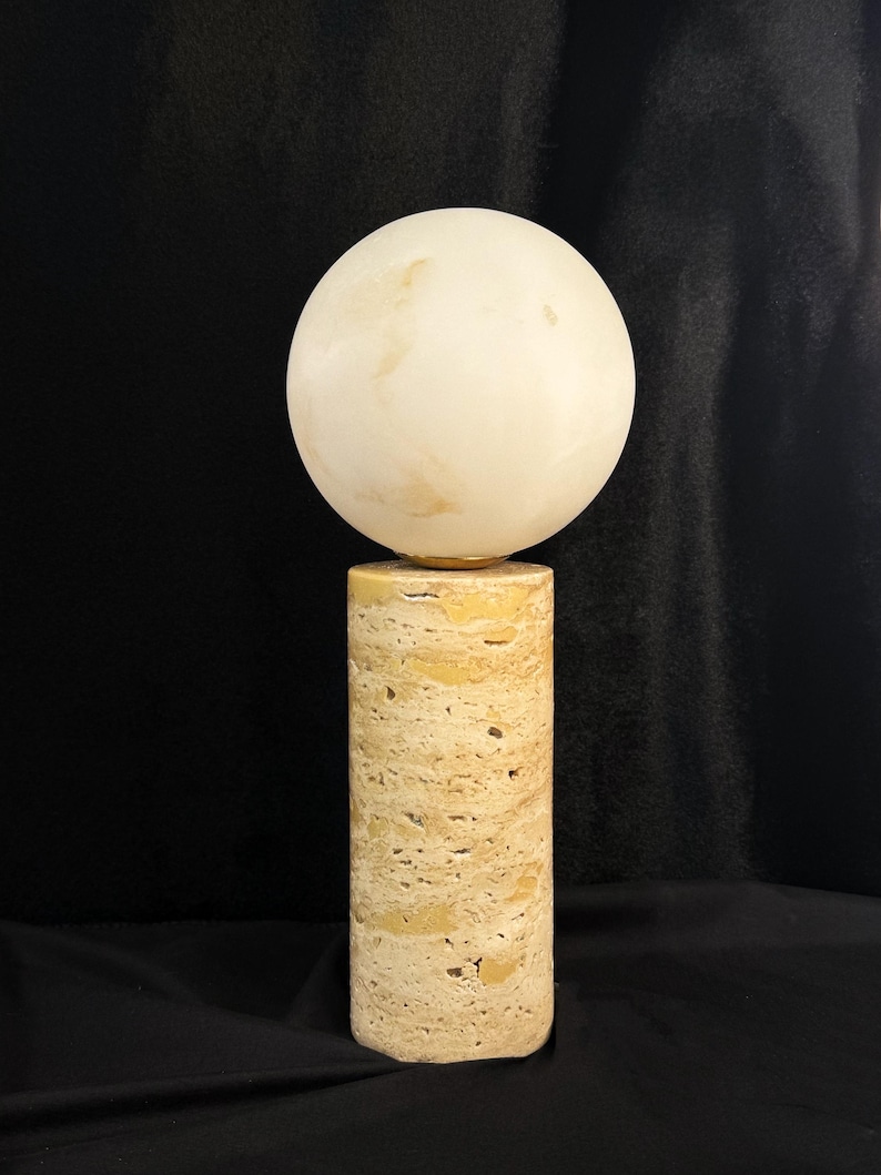 May include: A table lamp featuring a cylindrical base made of porous, beige travertine stone. The base supports a smooth, spherical, off-white globe shade that sits directly on top of the stone column, creating a balanced and geometric lighting fixture.