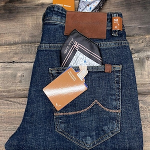 May include: Dark wash denim jeans with a brown leather patch and a pocket square. The jeans have a tag that reads "THE LUXURY PHILOSOPHY". The jeans are folded on a wooden surface.