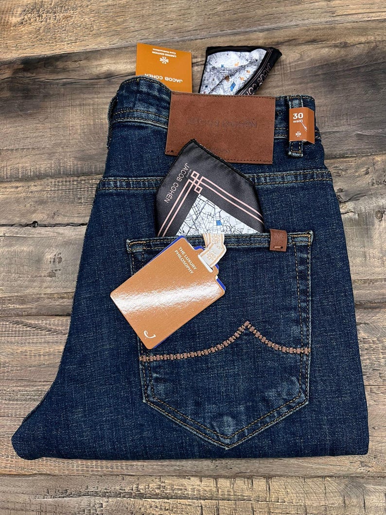 May include: Dark wash denim jeans with a brown leather patch and a pocket square. The jeans have a tag that reads "THE LUXURY PHILOSOPHY". The jeans are folded on a wooden surface.