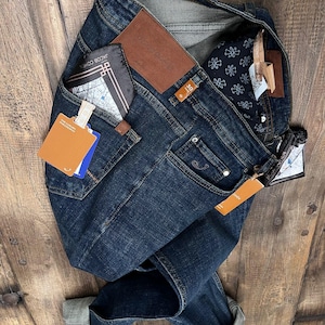 May include: Dark wash denim jeans with a brown leather patch and a folded cuff. The jeans have a pocket with a wallet and a tag. The jeans are on a wooden surface.
