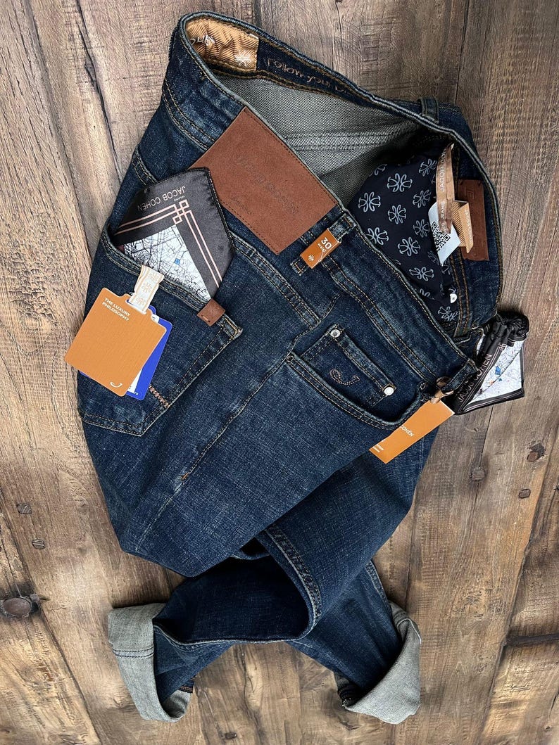 May include: Dark wash denim jeans with a brown leather patch and a folded cuff. The jeans have a pocket with a wallet and a tag. The jeans are on a wooden surface.