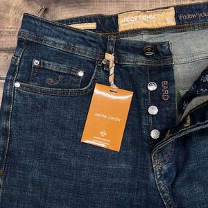 May include: Dark wash denim jeans with a button fly and a small coin pocket. The jeans have a branded tag that reads "JACOB COHEN" and "LIMITED EDITION". The jeans are new with tags.