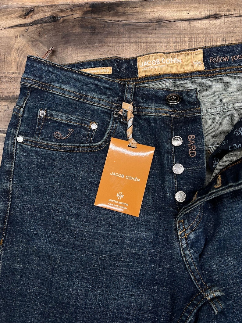 May include: Dark wash denim jeans with a button fly and a small coin pocket. The jeans have a branded tag that reads "JACOB COHEN" and "LIMITED EDITION". The jeans are new with tags.