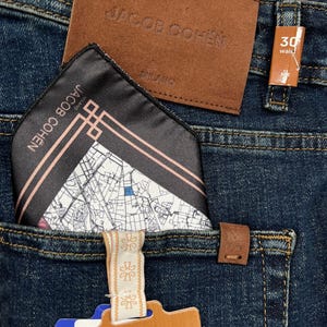 May include: Close-up of dark blue denim jeans with a brown leather patch and a pocket square. The pocket square is black with a map design and the brand name "JACOB COHEN". A tag reads "THE LUXURY PHILOSOPHY". Waist size is 30 inches.