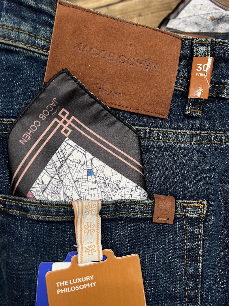 May include: Close-up of dark blue denim jeans with a brown leather patch and a pocket square. The pocket square is black with a map design and the brand name "JACOB COHEN". A tag reads "THE LUXURY PHILOSOPHY". Waist size is 30 inches.