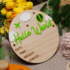 May include: A round wooden sign with the text "Hello World" in green letters. The sign features a hot air balloon, clouds, and stars. It also has spaces to write the date, time, weight, and length.
