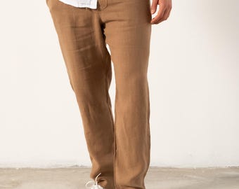 Può includere: Un paio di pantaloni di lino marroni con vita in cordoncino. I pantaloni sono indossati con una camicia di lino bianca e scarpe da ginnastica bianche.
