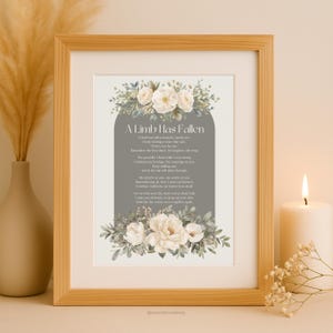 Puede incluir: Una impresión enmarcada con el texto "A Limb Has Fallen" rodeada de arreglos florales. La impresión está en un marco de madera clara, con una vela encendida y un jarrón en el fondo. El texto está en una fuente decorativa.