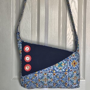 May include: A blue and white patterned crossbody bag with a navy blue triangular accent and three red buttons. The bag has a long strap and is made of fabric. The pattern features blue, yellow, and white floral tile designs.