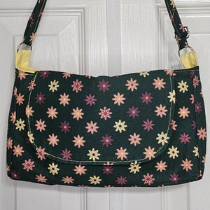 Cartera hecha a mano para adulto en color verde con flores naranjas, moradas y amarillas, correas para el hombro y tipo bandolera, bolsillo exterior y cierre de solapa.