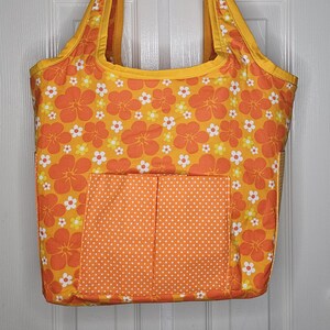 May include: A large orange tote bag with a floral print and a smaller orange pocket with white polka dots. The bag has two yellow handles.