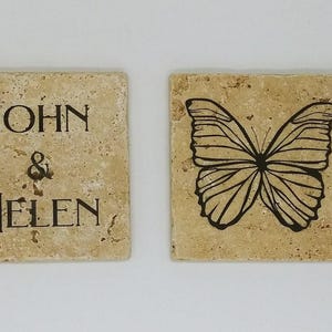 Könnte beinhalten: Zwei quadratische beigefarbene Untersetzer mit einer strukturierten Oberfläche. Ein Untersetzer trägt den Text "JOHN & HELEN" in schwarzen Buchstaben. Der andere Untersetzer hat eine schwarze Silhouette eines Schmetterlings.