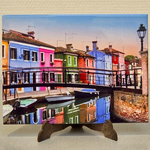 Puede incluir: Una hilera de casas coloridas con un puente sobre un canal. Las casas están pintadas en tonos de azul, rosa, verde y amarillo. Hay barcos en el canal y una farola en el lado derecho de la imagen.