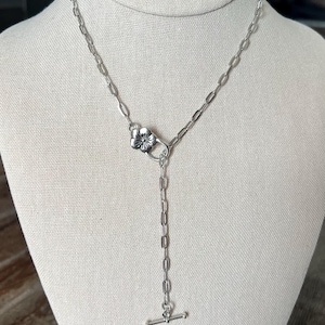 May include: Silver-toned necklace with a floral clasp and a vertical bar pendant. The necklace is displayed on a mannequin. The chain is made of oval links. The pendant has a T-bar closure.