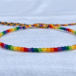 May include: A handmade rainbow friendship bracelet. The bracelet is made of colorful threads in a spectrum of colors, including red, orange, yellow, green, blue, and purple. The bracelet is adjustable and tied with a braided closure.