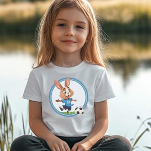 May include: White t-shirt with a blue circle graphic featuring a cartoon bunny wearing a blue soccer jersey with the number 10 on it. The bunny is kicking a soccer ball.
