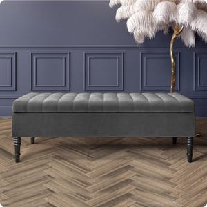 May include: A gray upholstered storage bench with a tufted top and dark wooden legs. The bench is rectangular and sits on a wooden floor with a herringbone pattern. The background features a dark blue wall with decorative panels.