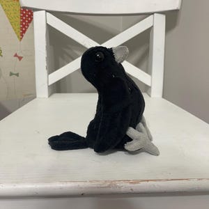 Puede incluir: Un juguete de peluche negro con detalles grises, posado sobre una silla de madera blanca. El pájaro tiene cola y pequeñas alas. La silla tiene respaldo en forma de X y está pintada de blanco. El fondo es una pared gris neutra.