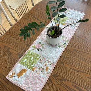 Peut inclure: Chemin de table matelassé avec un motif patchwork de carrés roses, blancs et verts. Les carrés présentent des motifs floraux, des chiens corgi et des pois. Une plante en pot repose sur le chemin de table, posé sur une table en bois.