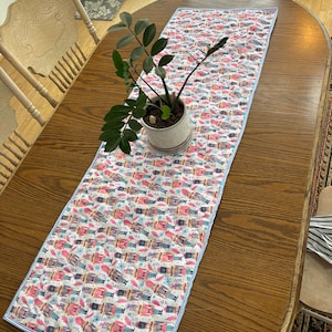 May include: A rectangular table runner with a repeating pattern of nutcrackers, snowflakes, and pink and blue accents on a white background. The runner is bordered with a light blue trim. A potted plant sits on the runner.