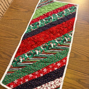 Peut inclure: Chemin de table de Noël matelassé avec une bordure blanche. Il présente des rayures diagonales en tissus rouge, vert et bleu marine. Certaines rayures ont des motifs de vacances comme des flocons de neige, des cannes en bonbon et des corgis avec des chapeaux de Père Noël.