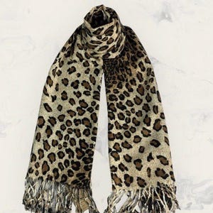 May include: A leopard print scarf with a beige background and brown spots. The scarf has a fringed edge and is draped. This fashion accessory measures approximately 180cm in length.