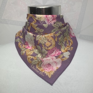 May include: A square, purple scarf featuring a floral pattern with pink and white roses, and gold and green foliage. The scarf is folded and draped, with the words "Christian Dior" printed on the corner.