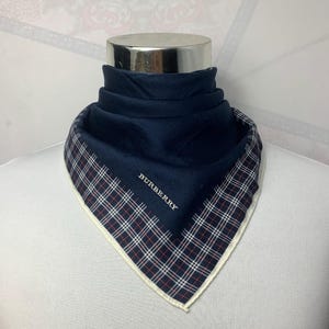 May include: A navy blue scarf with a plaid border in red, white, and black. The scarf is folded and draped over a mannequin head. The word "BURBERRY" is printed in white on the scarf.
