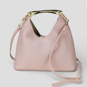 Könnte beinhalten: Eine hellrosa Handtasche mit einem goldfarbenen Griff und einem abnehmbaren Schulterriemen. Die Tasche hat ein glattes, strukturiertes Aussehen und ein schlichtes, elegantes Design. Der Riemen ist verstellbar.