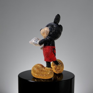 Side profile view of the "MICKEY LOVE" Pop Art statue, showing the figurine from the side as it stands on a black cylindrical pedestal. The collectible sculpture is covered in high-quality, hand-applied black, gold, and red glass crystals. The details of Mickey Mouse's shorts and shoes, and the shimmering texture of the rhinestones are clearly visible, highlighting the luxurious and unique aesthetic of this handmade art piece.