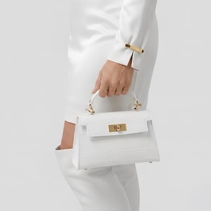May include: A white handbag with a crocodile-embossed texture, featuring a structured rectangular shape, a top handle, and gold-toned metal hardware including a rectangular turn-lock closure and side attachments for the handle.