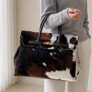 Puede incluir: Un bolso grande con estampado de piel de vaca con parches de pelo negro, marrón y blanco. El bolso incluye dos asas superiores de cuero negro, una correa horizontal de cuero negro en la parte delantera y un cierre de metal dorado.
