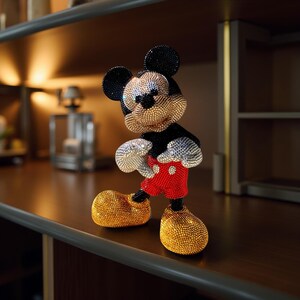 Stunning handmade beaded Mickey Mouse figurine. The collectible statuette stands on a wooden shelf, covered in thousands of tiny, sparkling beads. His head and torso are black and gold, his iconic shorts are red, and his oversized shoes are bright yellow. The figure's texture is highly detailed, catching the light beautifully. Ideal for Disney fans and collectors of unique, handcrafted art.