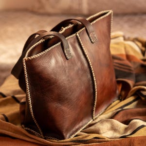 Puede incluir: Un bolso tote de cuero marrón con costuras blancas. El bolso tiene dos asas y un cierre de cremallera.