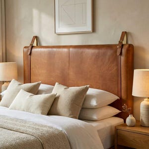 May include: A tan leather headboard with brass buckles and straps. The bed is made with white and beige pillows and a beige blanket. Two lamps with beige shades are on either side of the bed.