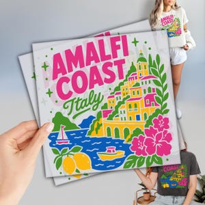 May include: A white t-shirt featuring a colorful graphic print of the Amalfi Coast in Italy. The design includes bright pink text reading AMALFI COAST, green foliage, yellow buildings, a blue sea with a sailboat, a small boat, and yellow lemons.