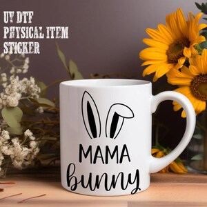 A white mug with a black bunny ear design and the text "MAMA bunny" printed on it, placed on a wooden surface next to a bouquet of yellow sunflowers.