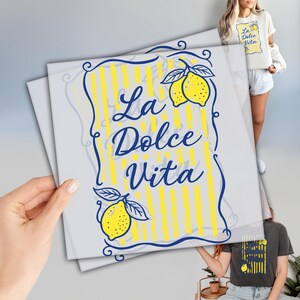 May include: A graphic design featuring the text "La Dolce Vita" in blue script, framed by a blue border and yellow stripes, with lemon illustrations. The design is displayed on a white background and is also printed on a t-shirt.