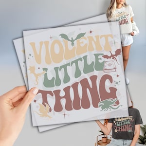 May include: A white graphic tee with the text "VIOLENT LITTLE THING" in a retro font. The text is surrounded by stars and images of dragons and a book.
