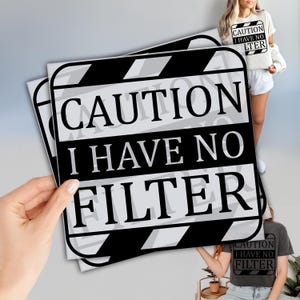 May include: A black and white sign with the text "CAUTION I HAVE NO FILTER" in a bold, sans-serif font. The sign has a film clapperboard design. The image also shows a person wearing a t-shirt with the same text.