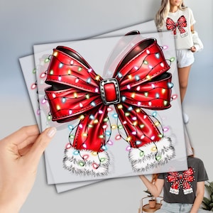 May include: A red bow design with a silver buckle and white fur trim is featured on a square print. The bow is adorned with colorful string lights. The image is held by a hand, and a person is wearing a shirt with the same bow design.