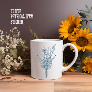A white mug with a blue floral design sits on a wooden surface, surrounded by yellow sunflowers and white flowers.