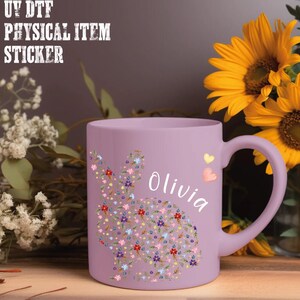 May include: A lavender ceramic mug featuring a floral bunny design and the name "Olivia" in white script. The bunny is composed of colorful flowers. Two small hearts are next to the name. The mug is next to sunflowers.